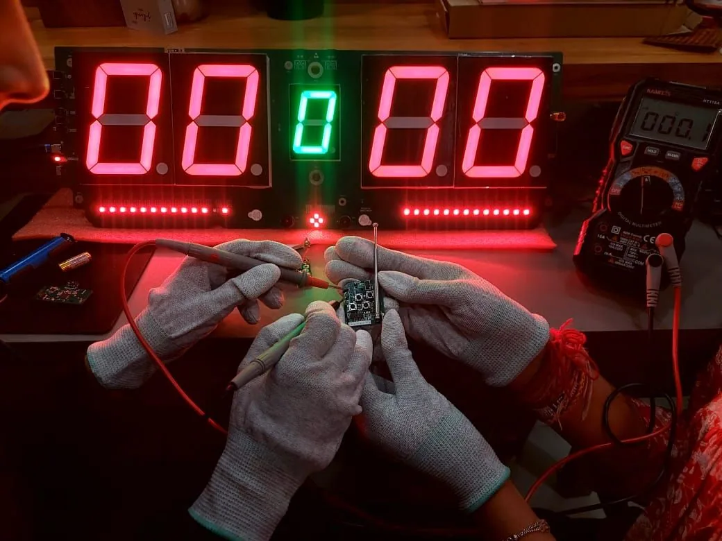 Engineer in ESD gloves probing the remote PCB with multimeter leads in front of the lit pickleball scoreboard prototype
